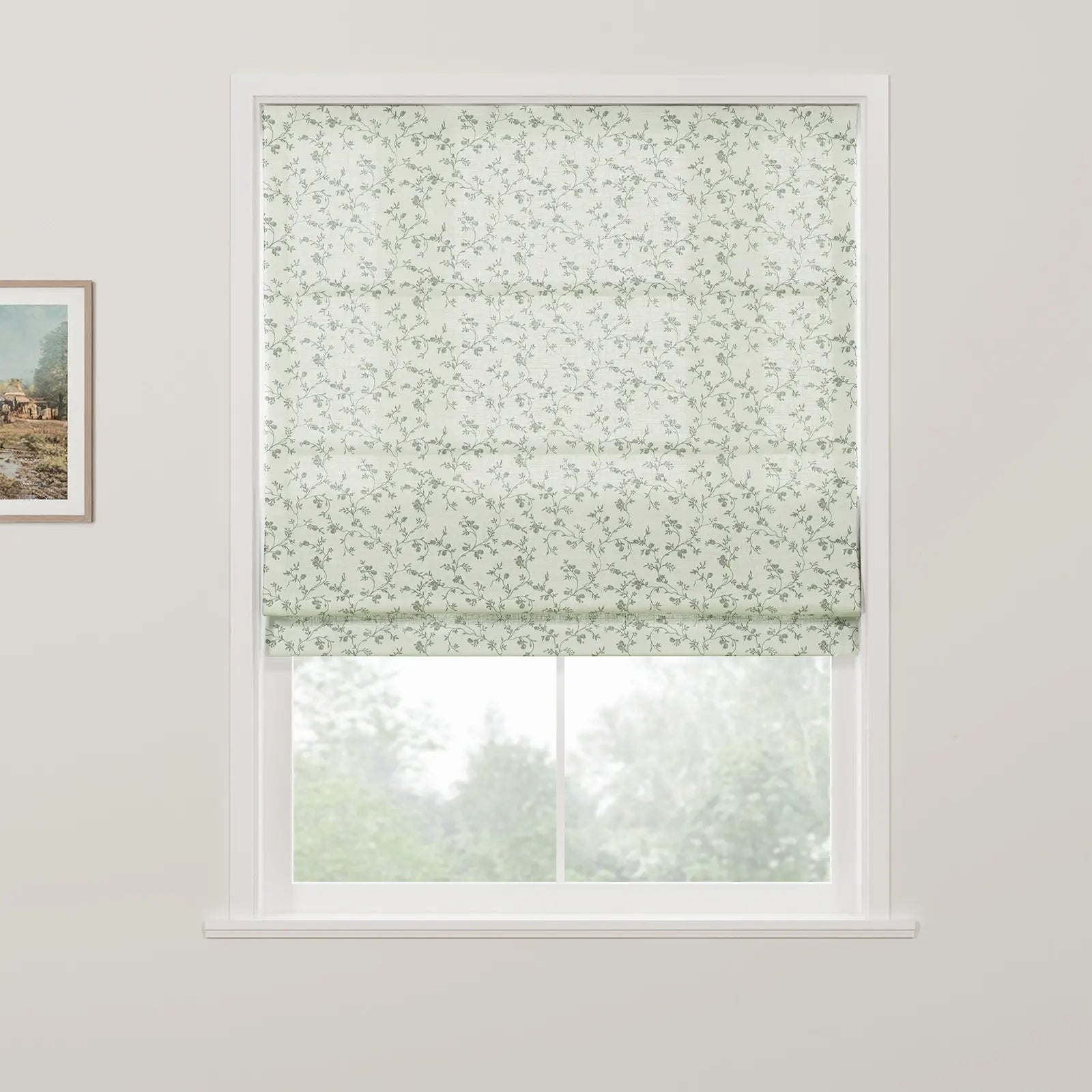 Partially lowered light green Roman blind with a subtle greyish-green floral and leaf pattern on a white framed window overlooking blurred green trees, with a small framed landscape painting visible on the left wall