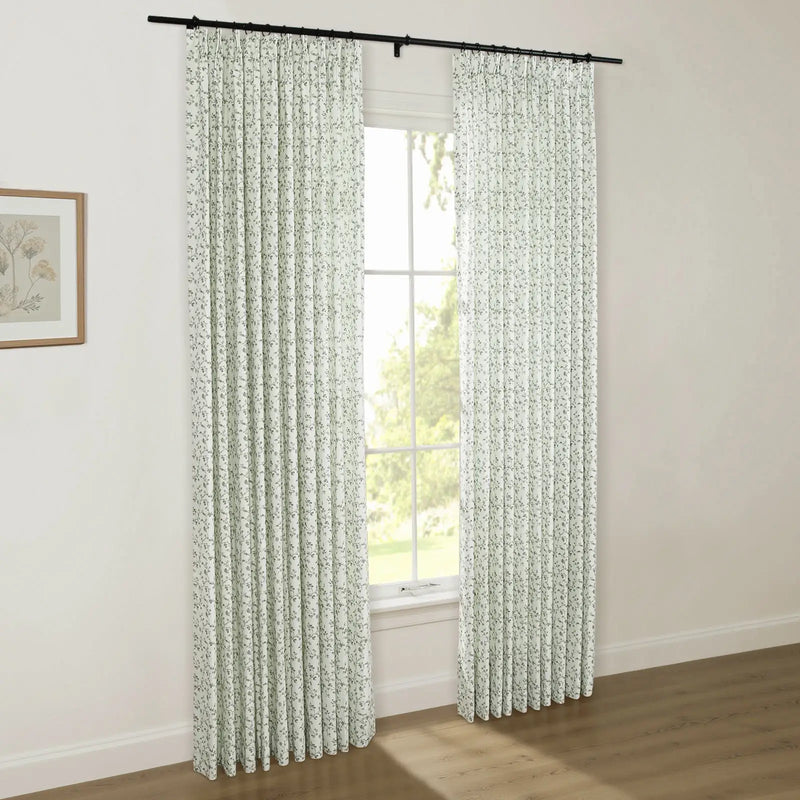 Photograph of a bright room with a window featuring white pleated curtains adorned with a small green botanical print, hung on a black rod, alongside a natural wood framed botanical art print on the wall and a view of lush green trees outside