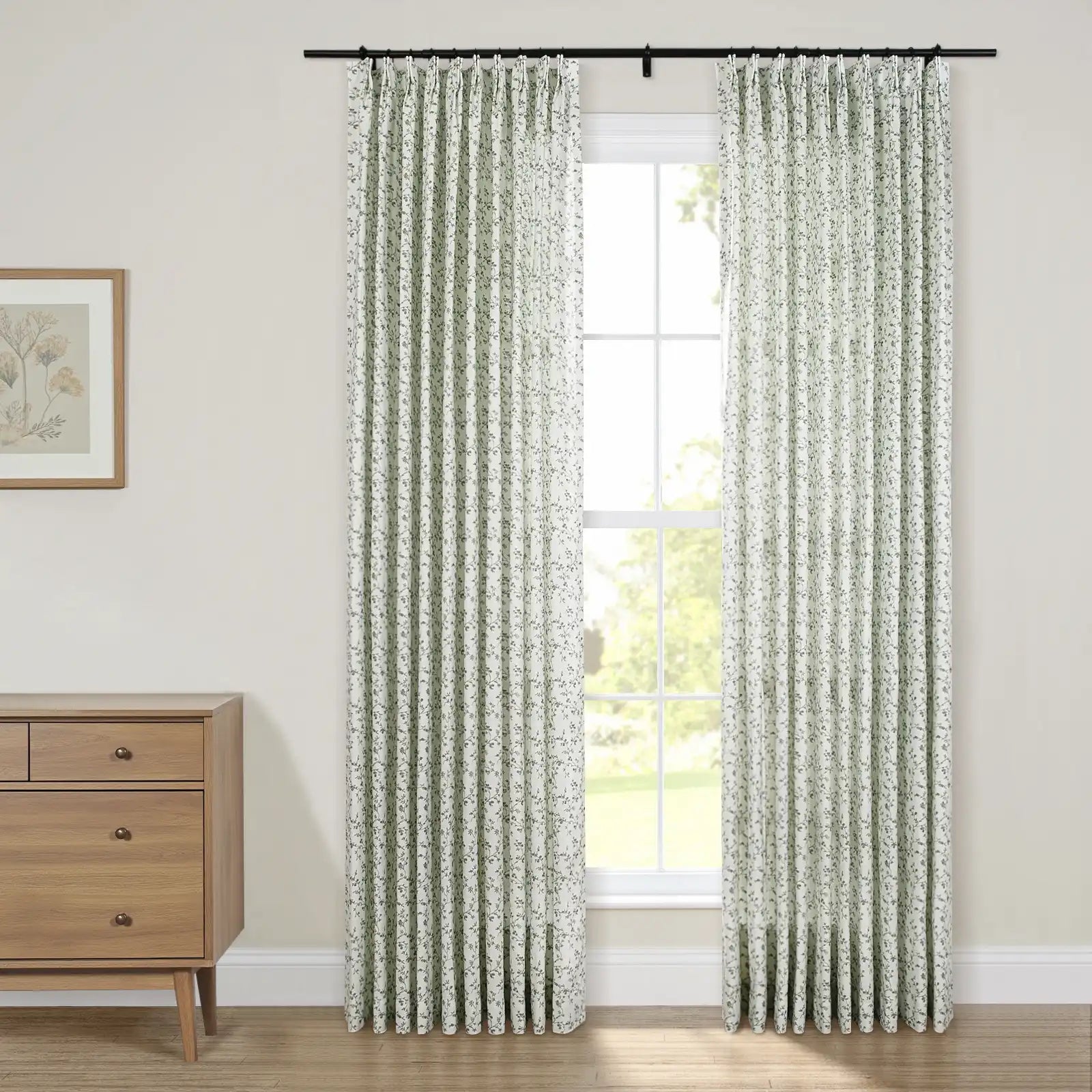 Interior view of a room with a large window adorned by white and green floral pinch-pleat curtains on a black rod, alongside a light wood mid-century modern dresser with bronze handles and a framed botanical art print