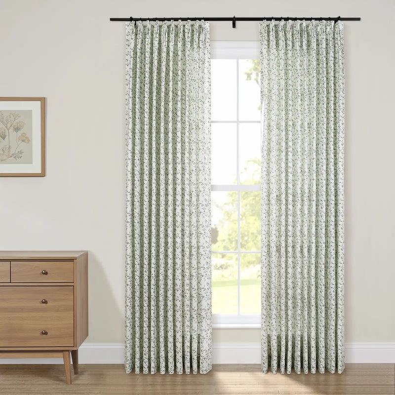 Interior view of a room with a large window adorned by white and green floral pinch-pleat curtains on a black rod, alongside a light wood mid-century modern dresser with bronze handles and a framed botanical art print
