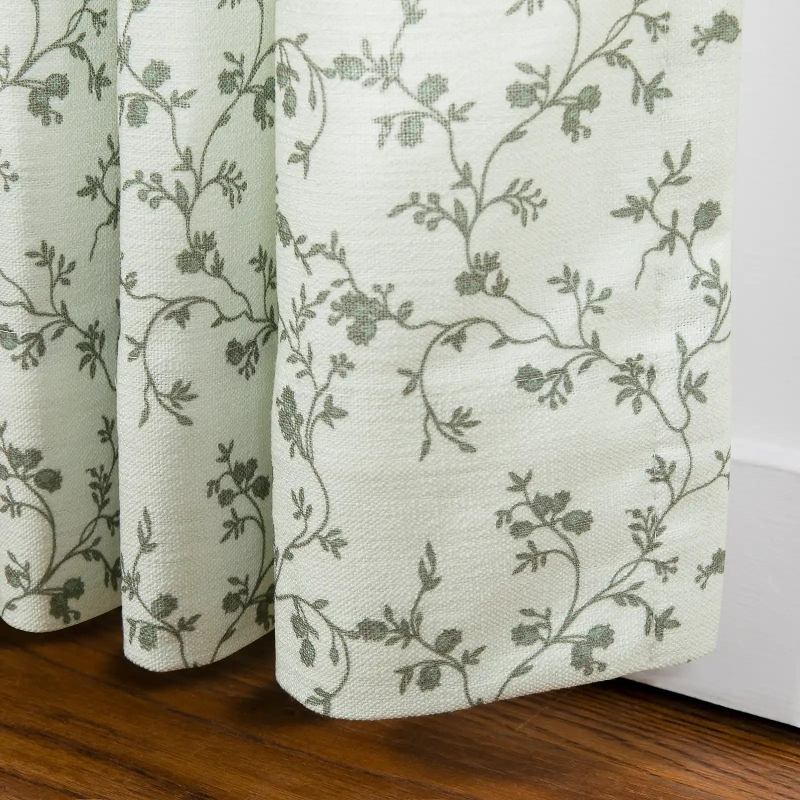 Fabric light green textured textile featuring a delicate grey-green floral vine pattern draped near a warm wooden floor and white baseboard