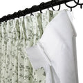 A light green fabric curtain with a delicate grey floral vine pattern layered behind a sheer white curtain, both suspended from a black metal rod using black rings and white plastic hooks