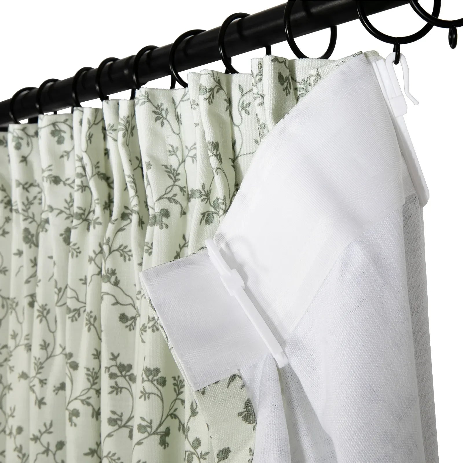 A light green fabric curtain with a delicate grey floral vine pattern layered behind a sheer white curtain, both suspended from a black metal rod using black rings and white plastic hooks