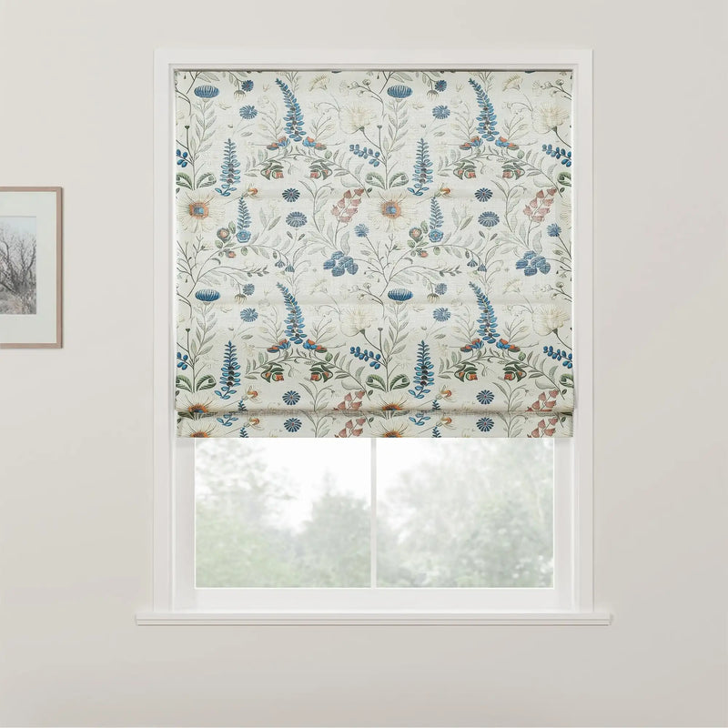 Textile Roman blind featuring a delicate floral pattern with blue lupine-like spikes, creamy white blossoms with orange centers, and small red flowers, set in a bright window against a backdrop of blurred green trees