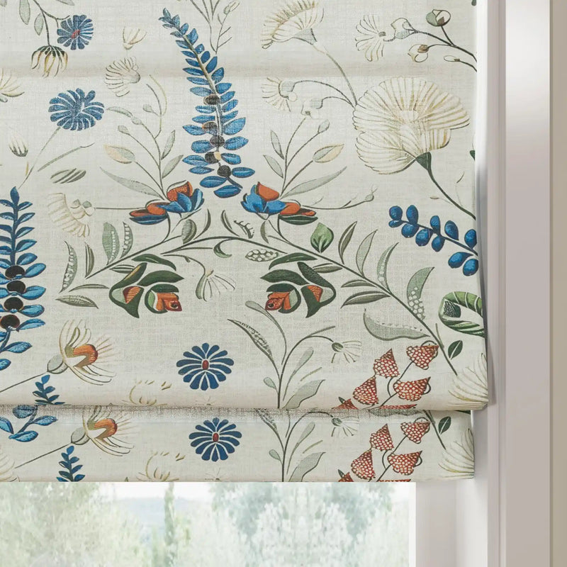 A floral patterned fabric Roman blind showcasing a chintz-like design with vibrant blue fern leaves, orange and blue bell flowers, and reddish berry clusters on a textured cream background at an indoor window