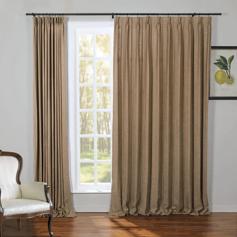 Beige textured drapes on a black rod frame a white paned window with a view of green trees in a room with a dark wood floor a cream antique armchair and a framed lemon botanical print