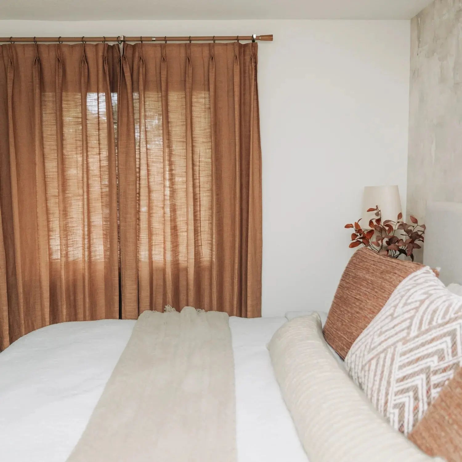 Bedroom interior featuring sheer terracotta curtains, white bedding with a beige runner, geometrically patterned pillows, and a bedside lamp with reddish autumn foliage