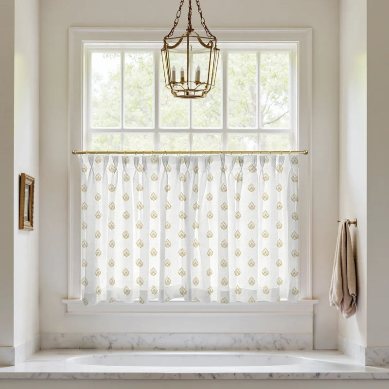 Bathroom window above a white built-in tub featuring a white valance curtain with a golden paisley pattern, a brass lantern chandelier, and a white marble surround