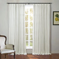 Photograph of a bright room featuring white linen-look pinch pleat curtains on a window overlooking sunlit trees an antique white upholstered armchair with carved wood and a framed lemon botanical print on the gray wall