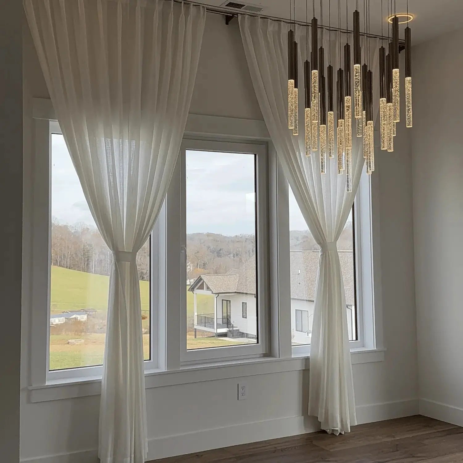 Modern interior room featuring large windows with sheer white curtains tied back, overlooking rolling green hills and modern homes, illuminated by a striking multi-pendant chandelier with clear bubble-filled glass tubes