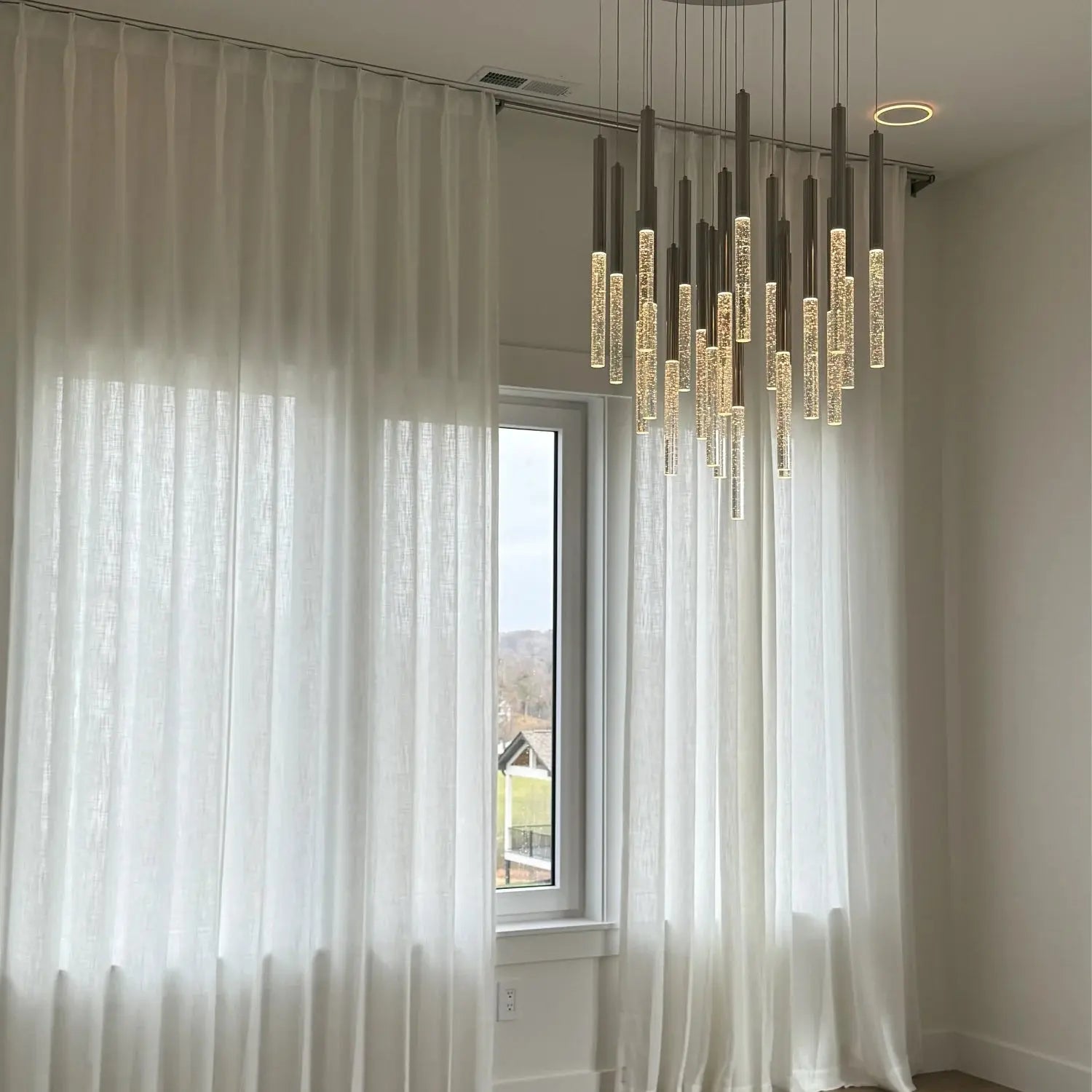 Modern room interior with full-length sheer white linen curtains framing a window overlooking suburban houses and hills, illuminated by a contemporary multi-pendant chandelier with glowing bubble-filled glass tubes