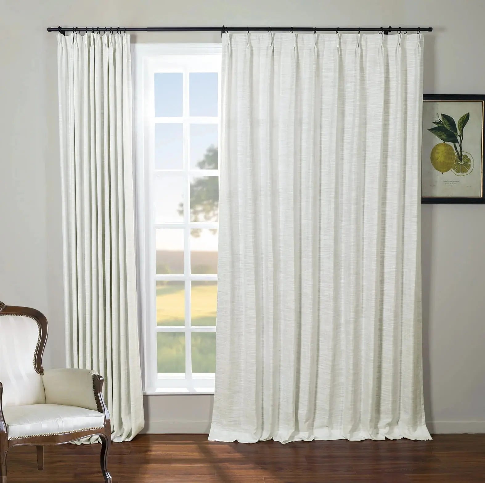 Bright room with white textured drapes on a black rod framing a grid-paned window looking out to a green field, featuring a classic wooden armchair with white upholstery and brass nailhead trim, and a framed lemon botanical print on the gray wall