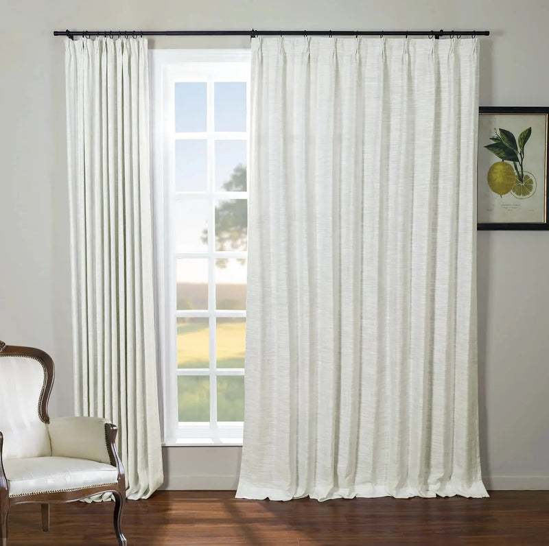Bright room with white textured drapes on a black rod framing a grid-paned window looking out to a green field, featuring a classic wooden armchair with white upholstery and brass nailhead trim, and a framed lemon botanical print on the gray wall