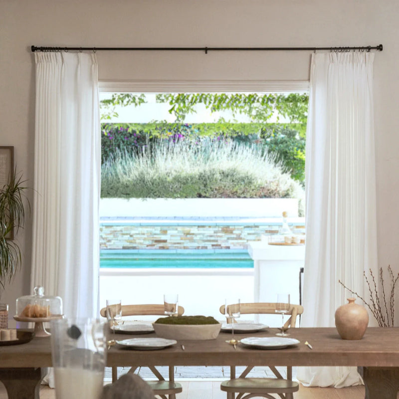 A rustic wooden dining table set with plates, glasses, and a moss centerpiece in a dining room, offering a view through white curtains to a blue swimming pool with colorful mosaic tiles and a lush garden of green and purple foliage