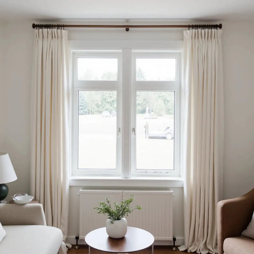 An interior shot of a modern living room window framed by cream pleated curtains on a dark rod, showcasing a white radiator below and a round wooden table with a white vase of green sprigs, looking out onto a distant field with a white utility vehicle