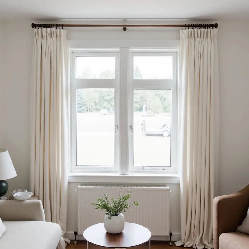 An interior shot of a modern living room window framed by cream pleated curtains on a dark rod, showcasing a white radiator below and a round wooden table with a white vase of green sprigs, looking out onto a distant field with a white utility vehicle