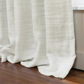 Photograph of textured white semi-sheer curtains elegantly pooling on a dark brown hardwood floor beside a white window frame