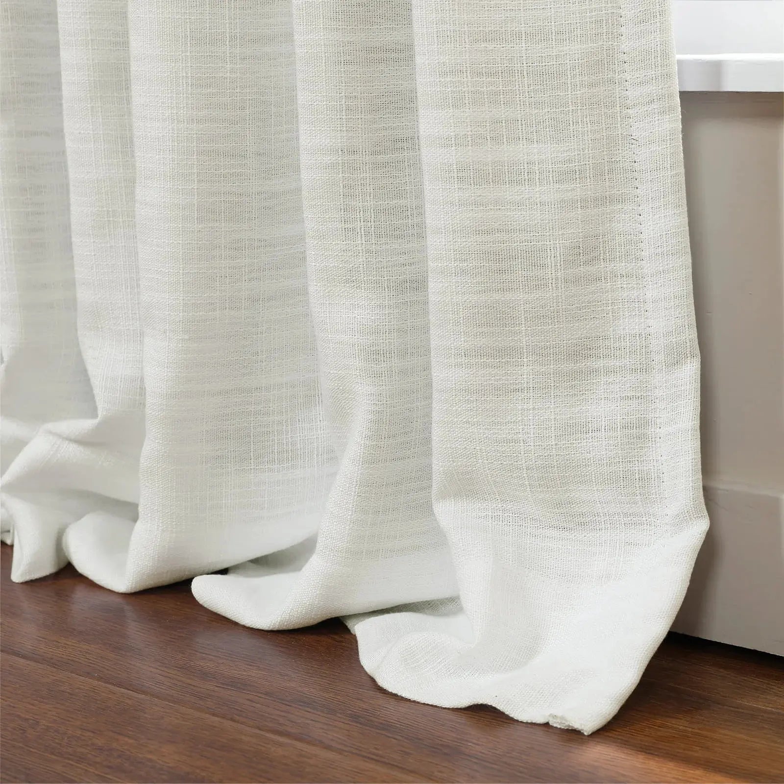 Photograph of textured white semi-sheer curtains elegantly pooling on a dark brown hardwood floor beside a white window frame
