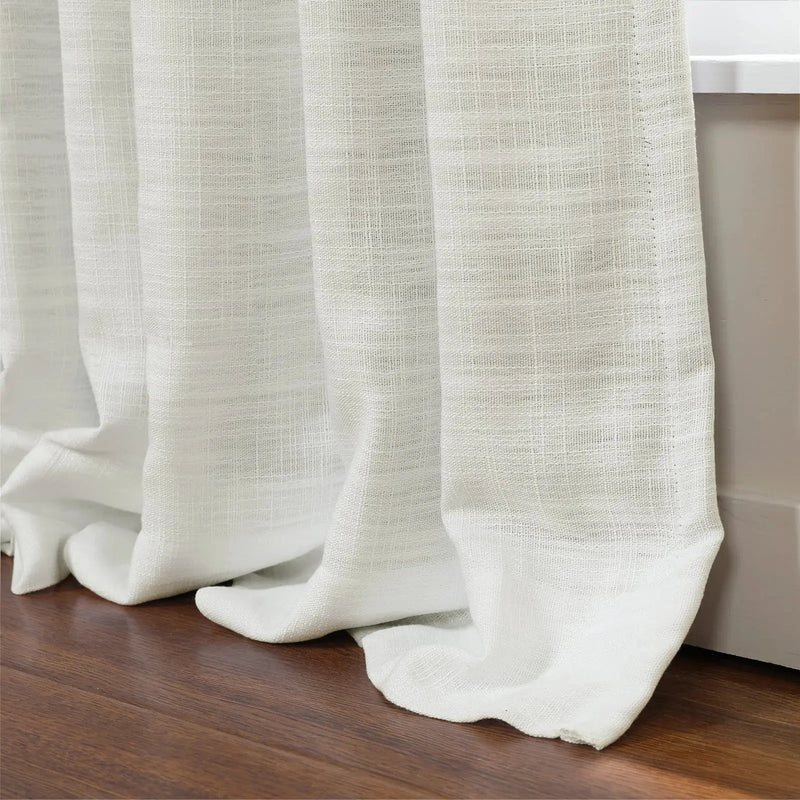 Photograph of textured white semi-sheer curtains elegantly pooling on a dark brown hardwood floor beside a white window frame