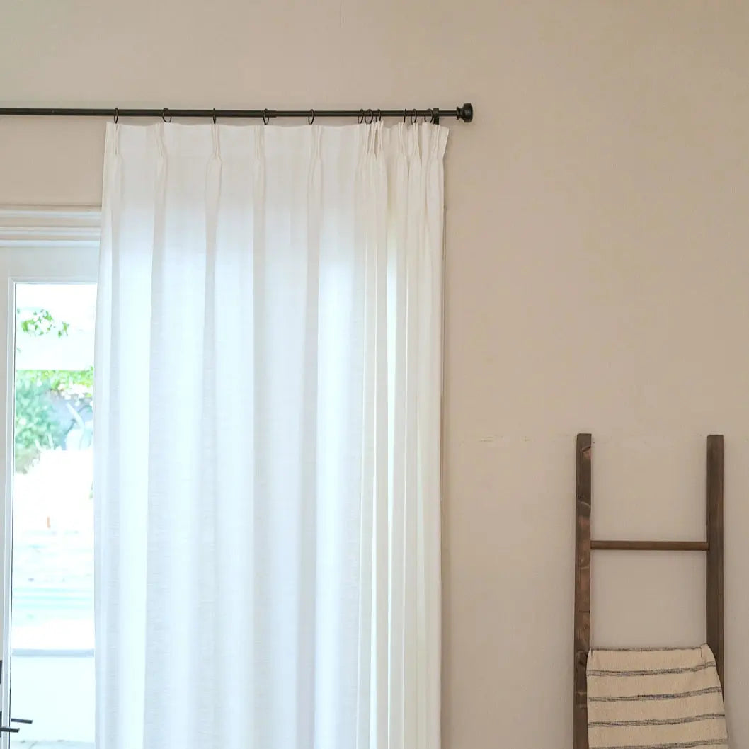 Photograph of a minimalist indoor scene with a sheer white curtain covering a glass door, a warm beige wall, and a dark wooden decorative ladder draped with a beige and dark gray striped towel
