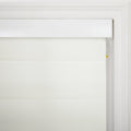 Photograph of a creamy white pleated window Blind with a textured fabric featuring a sleek white headrail and visible white control strings with a small yellow safety tag on the right, set against elegant white window molding