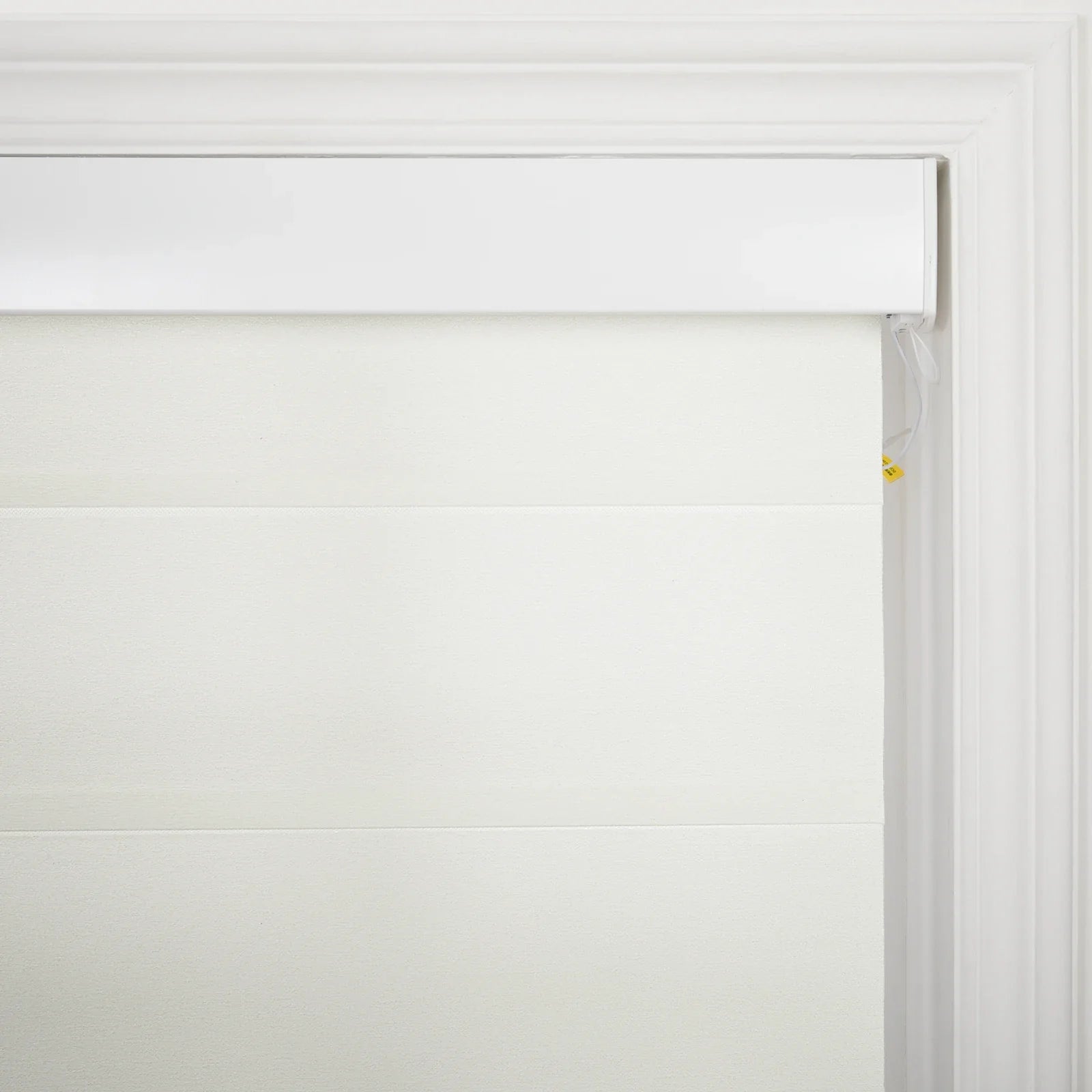 Photograph of a creamy white pleated window Blind with a textured fabric featuring a sleek white headrail and visible white control strings with a small yellow safety tag on the right, set against elegant white window molding