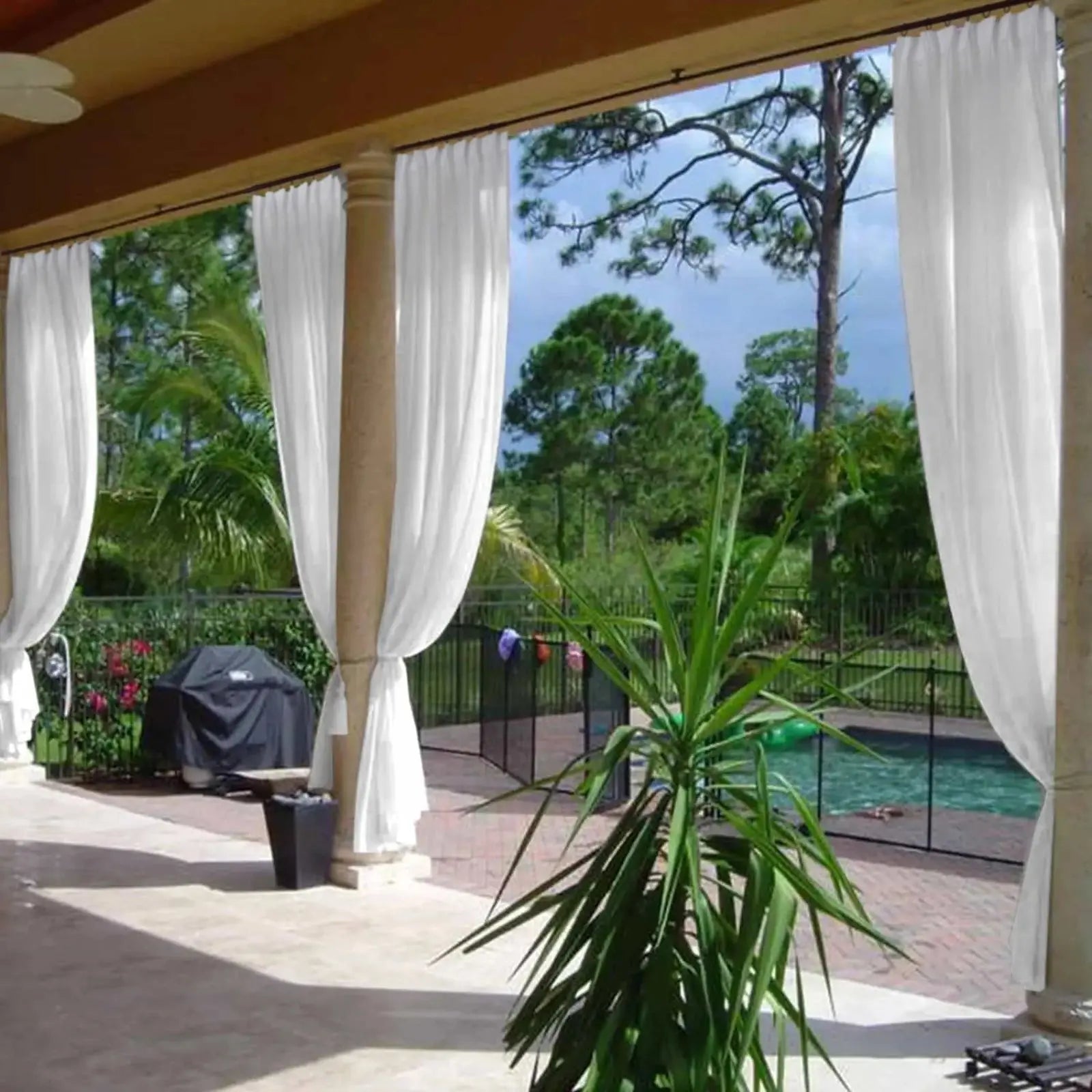 A resort-style outdoor patio with flowing white sheer curtains tied around substantial columns, revealing a fenced swimming pool with a green inflatable, a black covered barbecue grill, a vibrant green yucca plant, and a tropical landscape of palm and pine trees under a clear blue sky
