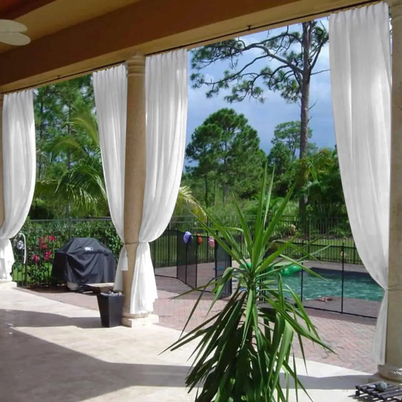A resort-style outdoor patio with flowing white sheer curtains tied around substantial columns, revealing a fenced swimming pool with a green inflatable, a black covered barbecue grill, a vibrant green yucca plant, and a tropical landscape of palm and pine trees under a clear blue sky