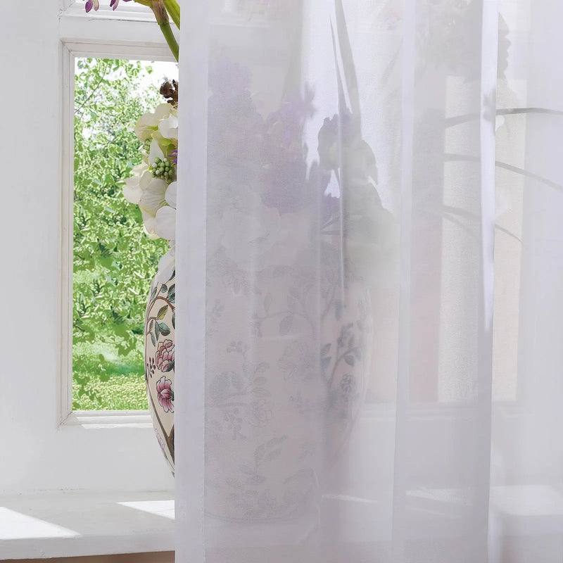Photography of a decorative ceramic vase featuring pink and green floral patterns filled with white hydrangeas and purple flowers on a white window sill, glimpsing a vibrant green garden through sheer white curtains