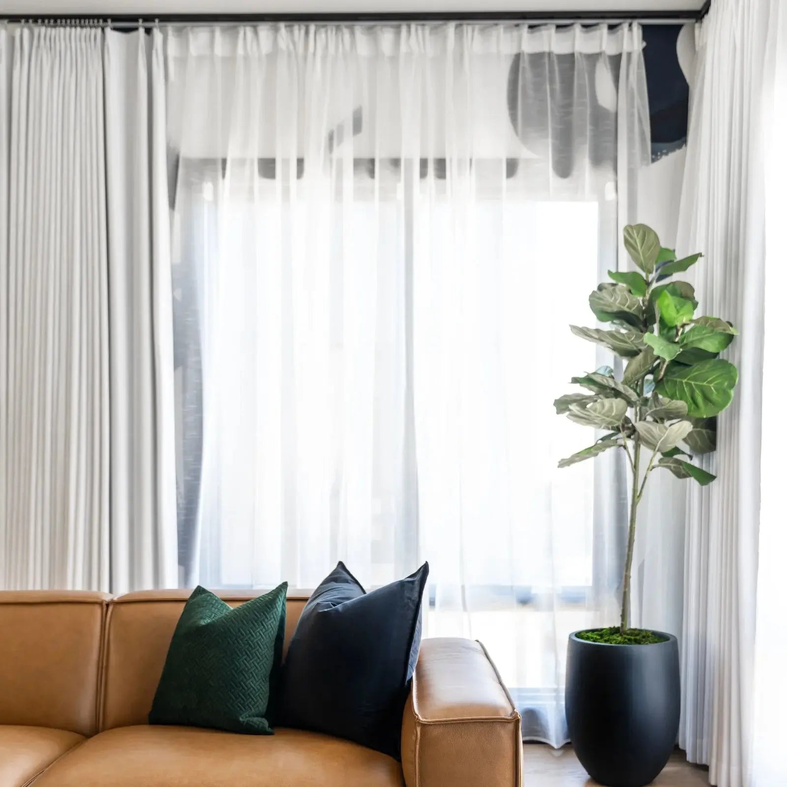 A modern living room interior with a tan leather sofa, vibrant green and black throw pillows, a tall fiddle-leaf fig plant in a sleek black planter, and sheer white curtains framing a window revealing subtle dark abstract wall art