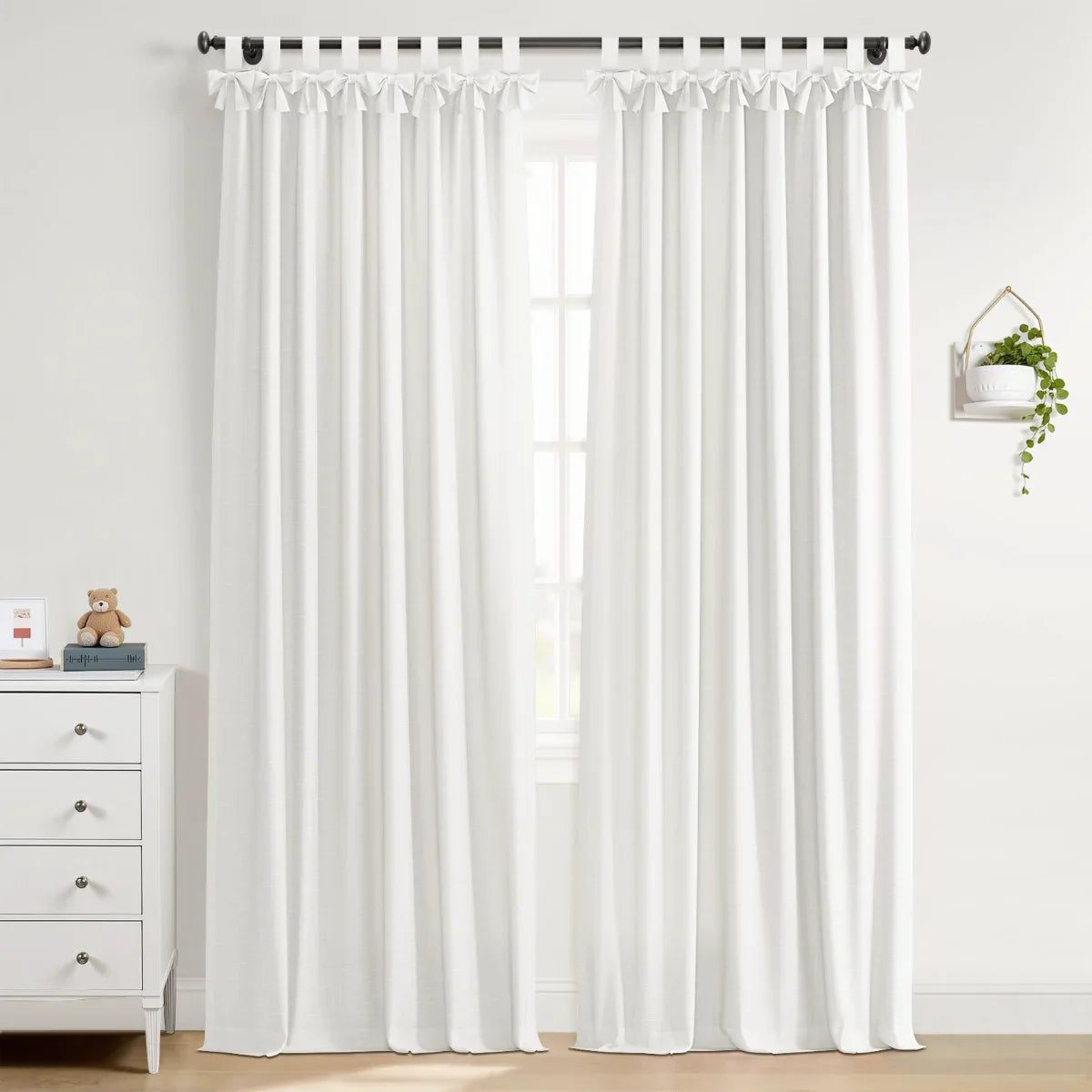 A pair of elegant white tie-top curtains on a black rod in a bright room featuring a white dresser with a teddy bear and framed art, plus a hanging green plant on a minimalist wall shelf