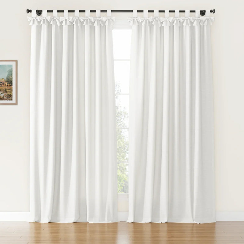 White linen texture curtains with decorative bow-tie tab tops hanging from a black rod in a sunlit room with hardwood floors, a visible outdoor green tree, and a small framed painting on the left wall