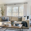 Photograph of a modern living room interior showcasing a neutral beige sectional sofa adorned with grey and patterned pillows and a dark knit blanket, a wooden coffee table holding a textured white bowl, a blue-green vase with dried flowers, and a lidded pot, all bathed in natural light from large windows framed by white sheer curtains