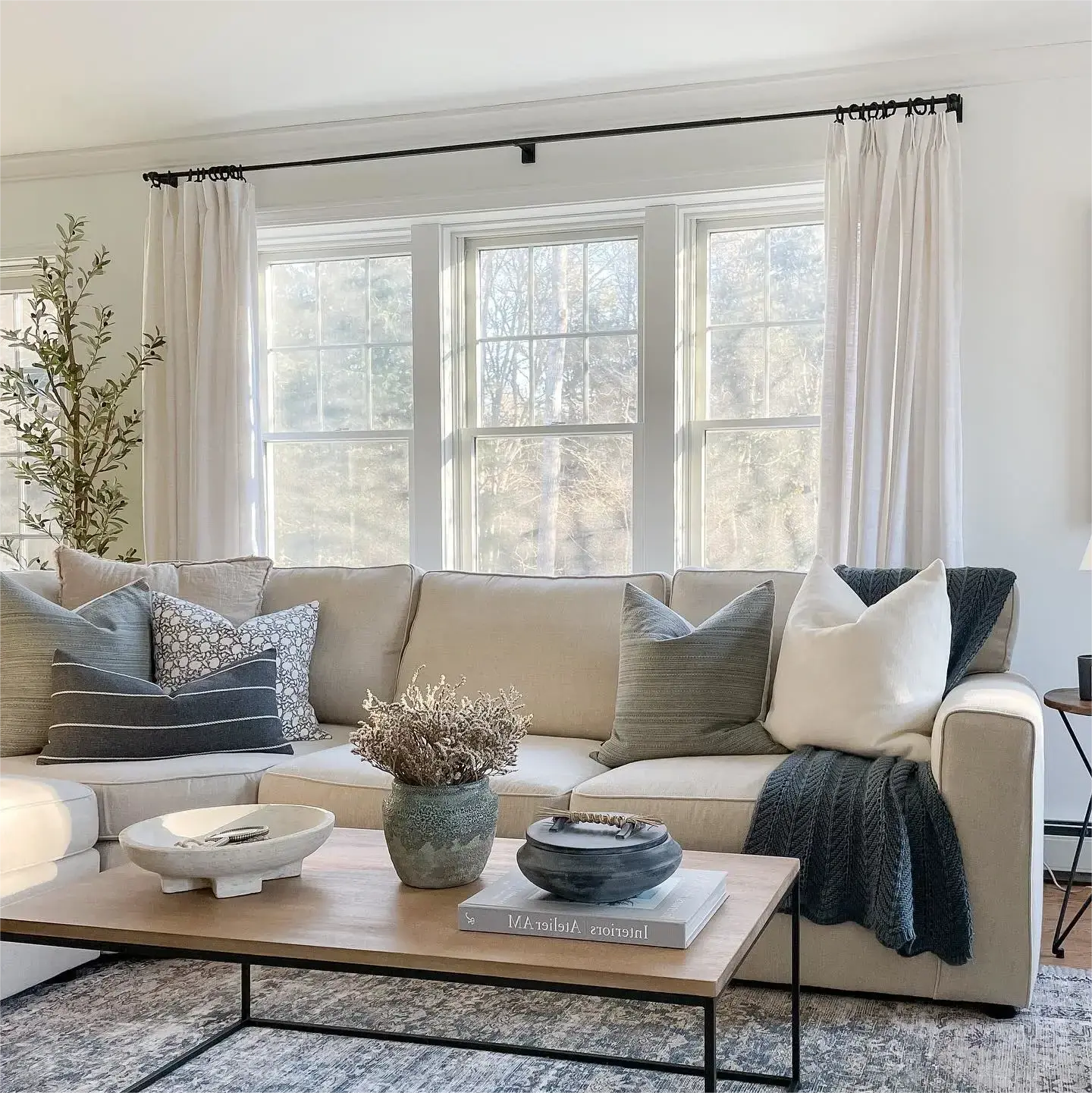 Photograph of a modern living room interior showcasing a neutral beige sectional sofa adorned with grey and patterned pillows and a dark knit blanket, a wooden coffee table holding a textured white bowl, a blue-green vase with dried flowers, and a lidded pot, all bathed in natural light from large windows framed by white sheer curtains