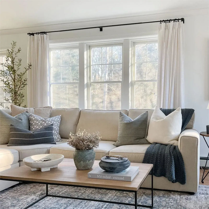 Photograph of a modern living room interior showcasing a neutral beige sectional sofa adorned with grey and patterned pillows and a dark knit blanket, a wooden coffee table holding a textured white bowl, a blue-green vase with dried flowers, and a lidded pot, all bathed in natural light from large windows framed by white sheer curtains