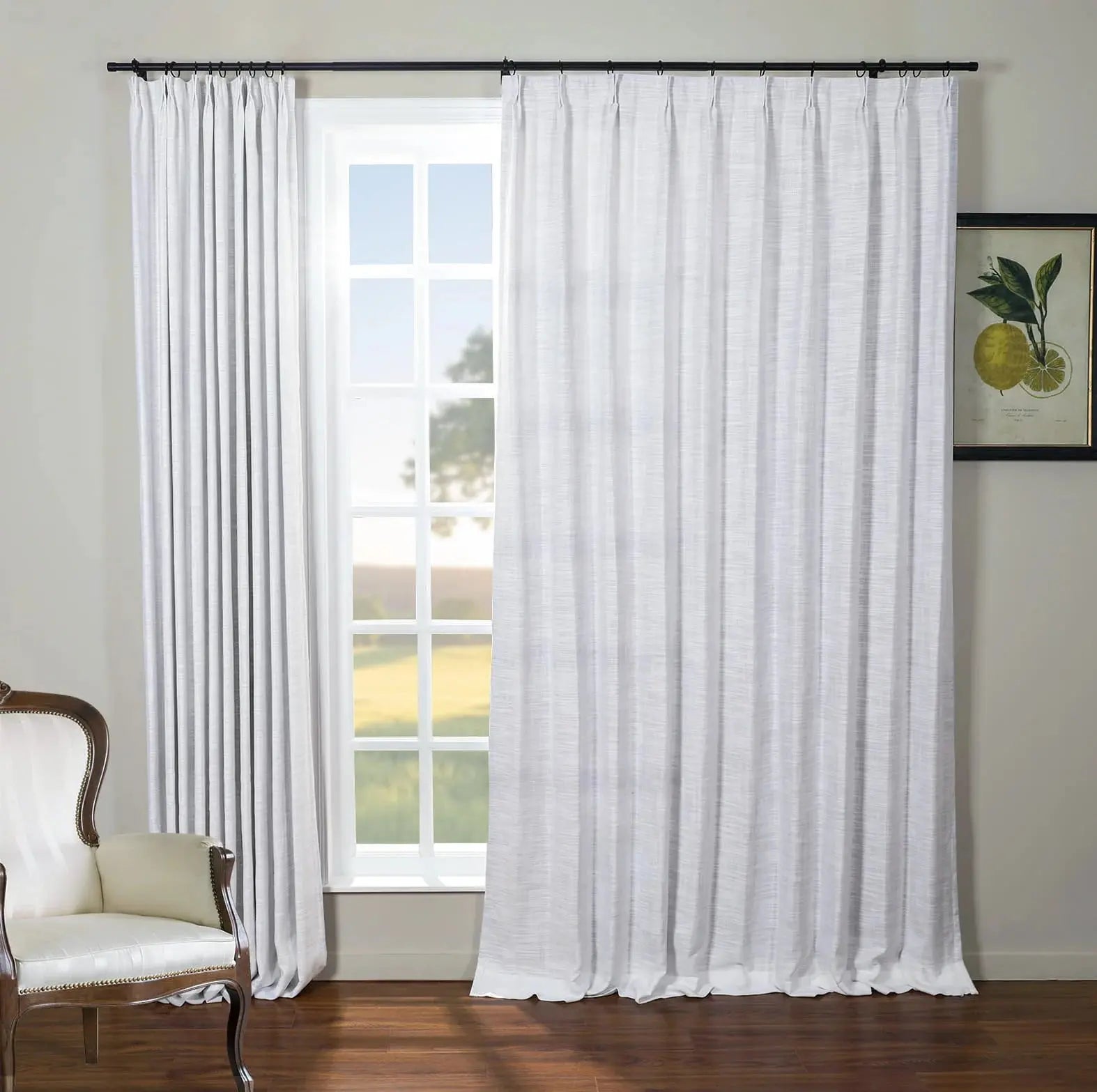 An interior room featuring a large window with white textured curtains, an elegant cream armchair with nailhead trim, and a framed botanical lemon print on the wall overlooking a sunny green field