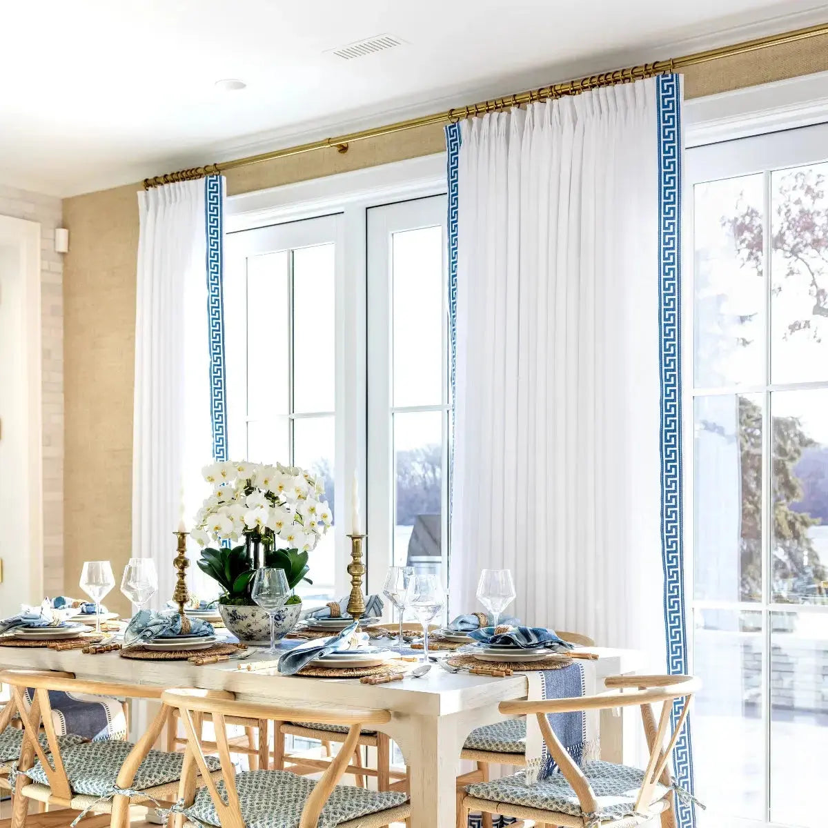A bright dining room with a long white table elegantly set with blue and white dishes, a lush white orchid centerpiece, and tall white drapes featuring bold blue Greek key trim framing a tranquil outdoor view
