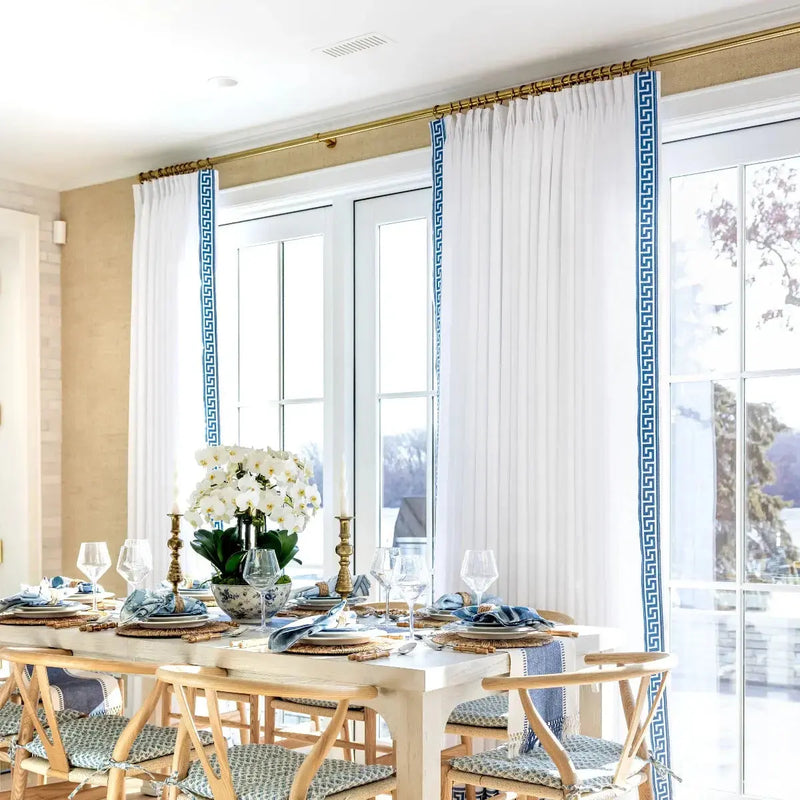 A bright dining room with a long white table elegantly set with blue and white dishes, a lush white orchid centerpiece, and tall white drapes featuring bold blue Greek key trim framing a tranquil outdoor view