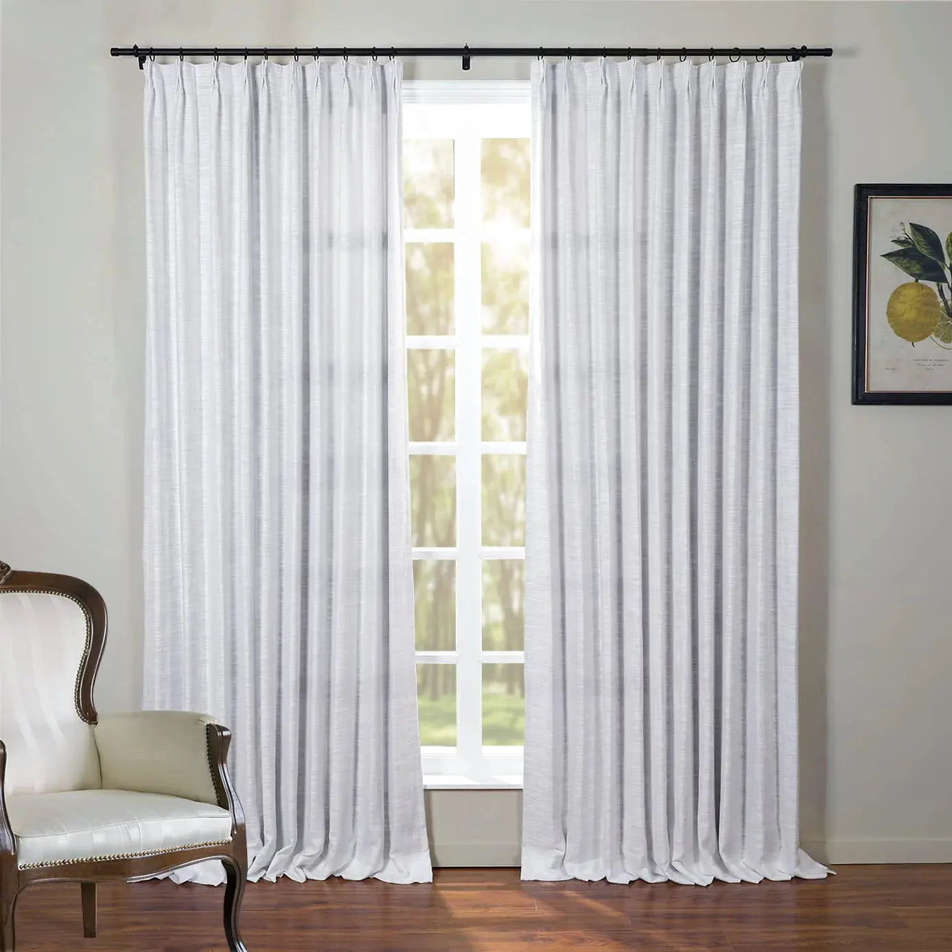 Photograph of white textured linen-blend curtains on a black rod, a classic wooden armchair with white upholstery and nailhead trim, and a framed lemon botanical print in a sunlit room overlooking green trees