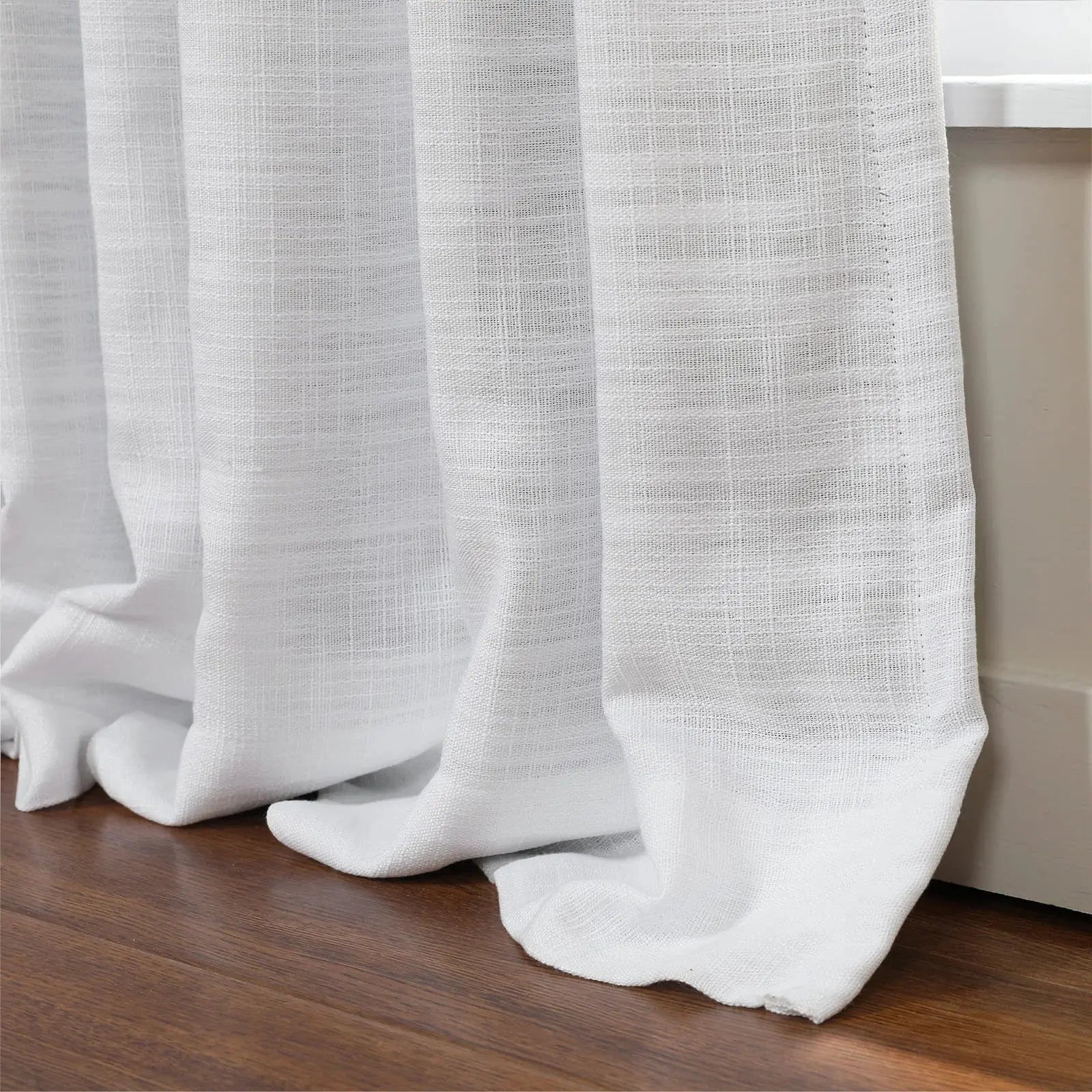 White sheer linen-look curtains with a visible textured weave pooling gracefully on a rich dark wood floor next to a crisp white window trim