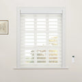 Photograph of a window with white motorized zebra Blinds featuring alternating sheer and opaque horizontal stripes, a wall-mounted remote control, and a framed botanical print, offering a view of vibrant autumnal trees outside
