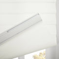 A modern white layered fabric window Blind with a visible headrail mechanism partially open revealing blurred green-yellow trees outside a window