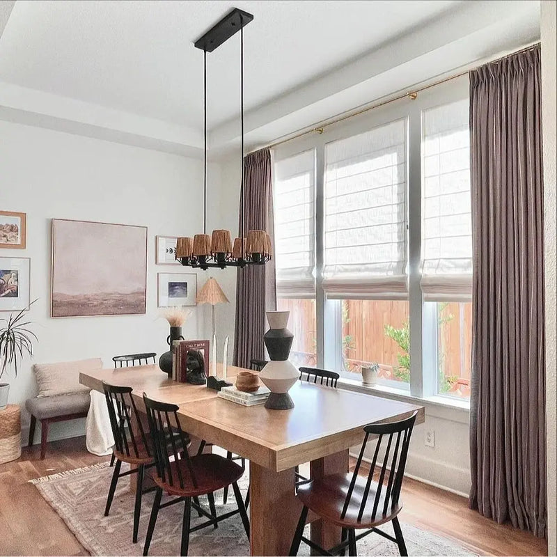 Photograph of a modern boho-chic dining room showcasing a light wood dining table with black chairs a linear black chandelier with six natural woven shades and a window wall featuring white Roman blinds flanked by mauve curtains
