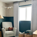 Interior photograph of a cozy nursery room featuring a white swan mobile, dark teal wainscoting below star-patterned wallpaper, a grey glider chair holding a fluffy brown highland cow plush, and black and white grid curtains