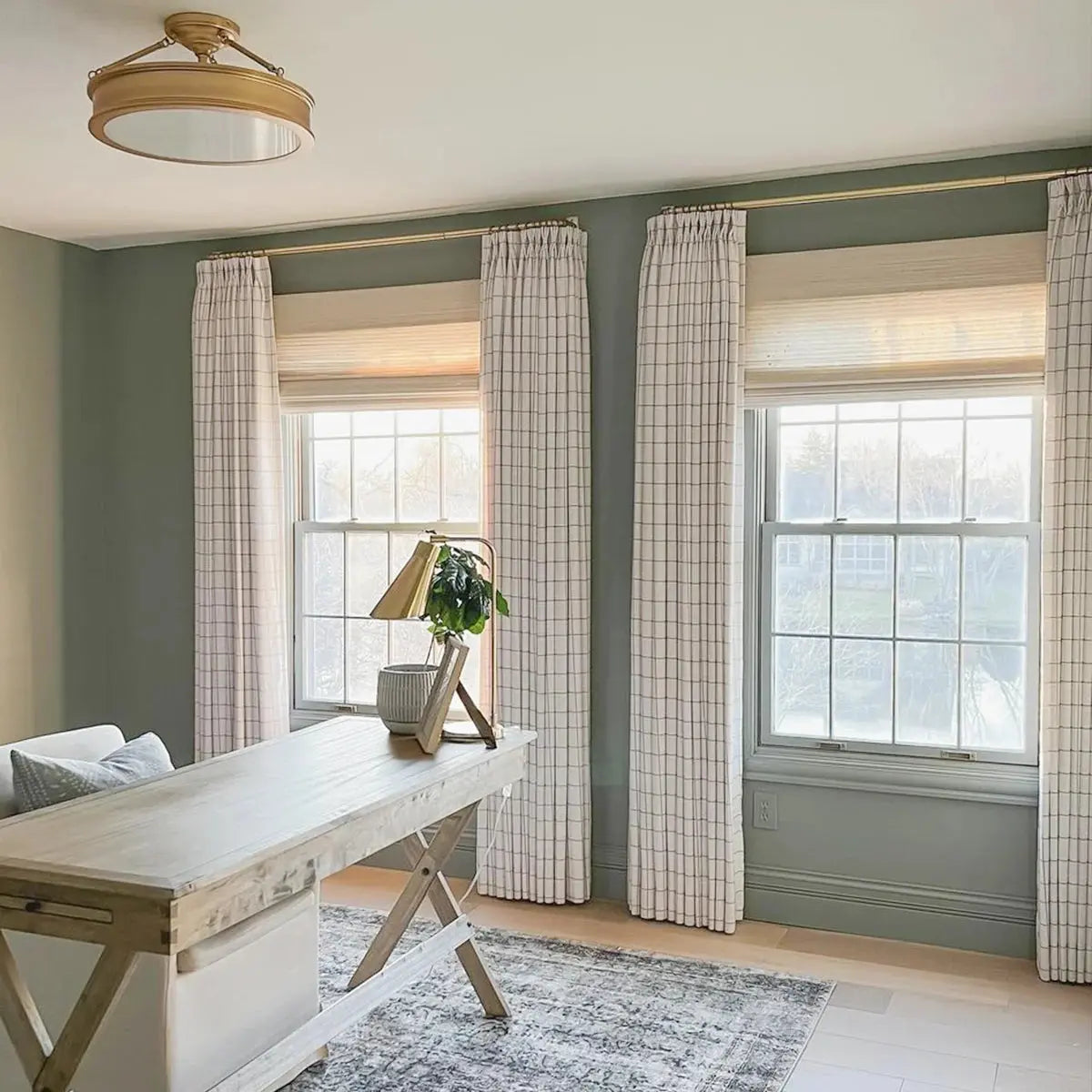 A bright home office with sage green walls featuring a light wood cross-leg desk, white windowpane curtains over natural woven blinds, and a brass ceiling light fixture