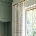 Interior view of a window featuring a white grid curtain on a brass rod next to deep green paneled cabinetry, with bright green foliage visible beyond