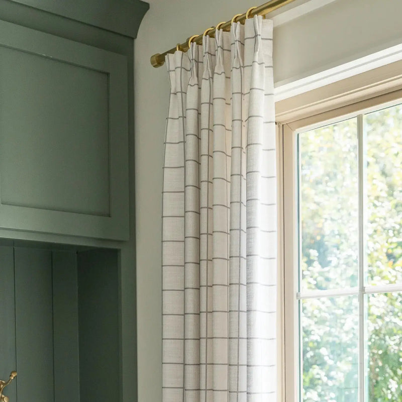 Interior view of a window featuring a white grid curtain on a brass rod next to deep green paneled cabinetry, with bright green foliage visible beyond