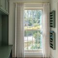 An interior view of a room with a large grid-paned window looking out to a green suburban yard, featuring light plaid curtains on a gold rod, sage green built-in cabinetry, and a matching wall-mounted sunglasses holder displaying vibrant blue and orange sunglasses
