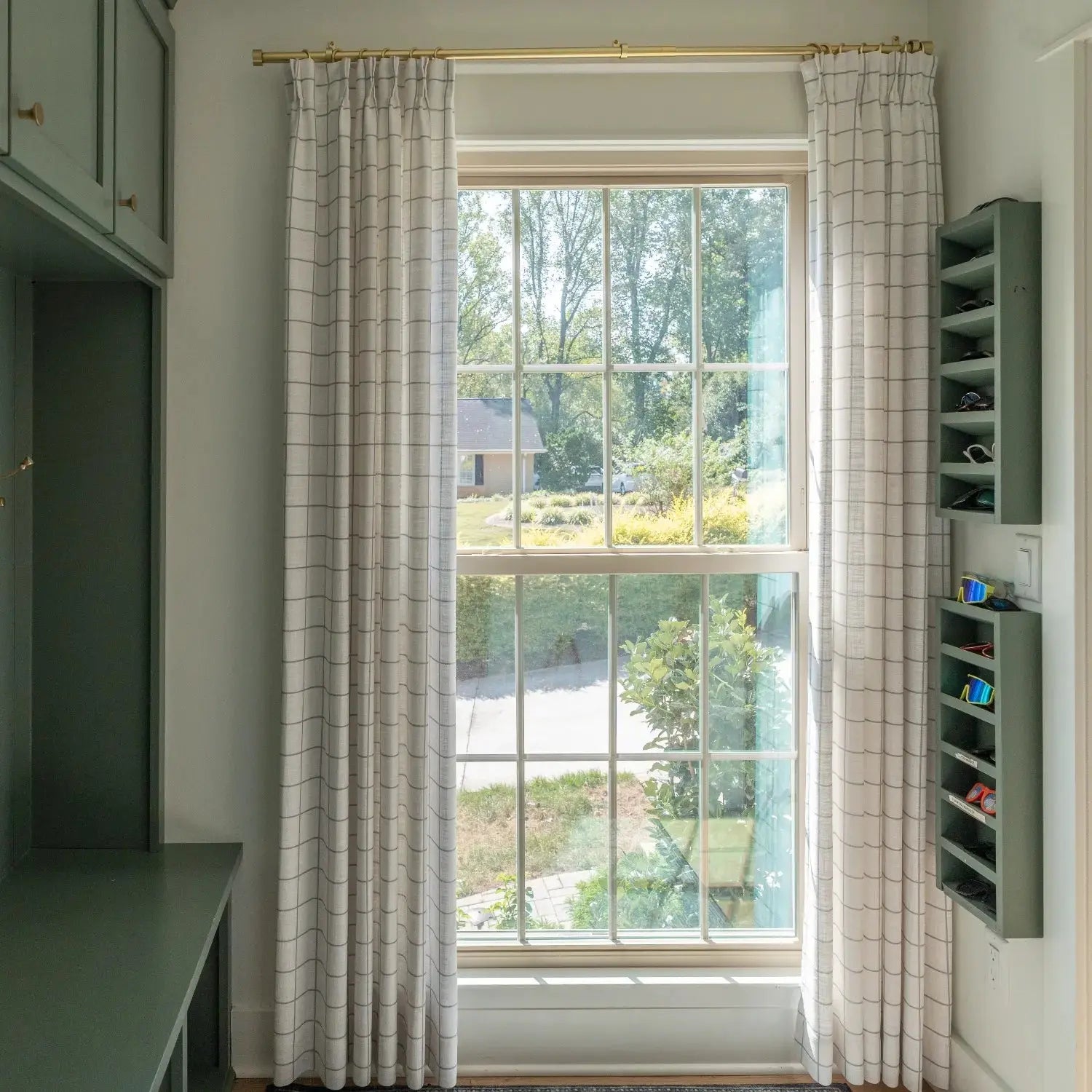 An interior view of a room with a large grid-paned window looking out to a green suburban yard, featuring light plaid curtains on a gold rod, sage green built-in cabinetry, and a matching wall-mounted sunglasses holder displaying vibrant blue and orange sunglasses