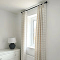 A bright bedroom interior featuring a window with beige grid-patterned curtains on a black rod, a white dresser with soccer ball knobs, and a dark grey textured lamp with a white shade